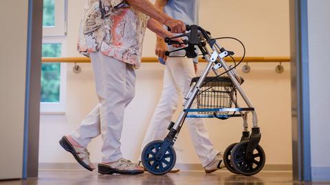 Im Vordergrund schiebt eine ältere Frau ihren Rollator durch einen Pflegeheim-Korridor. Im Hintergrund ist zu erkennen, dass sie von einer Pflegerin in einem blauen Kittel begleitet wird. 