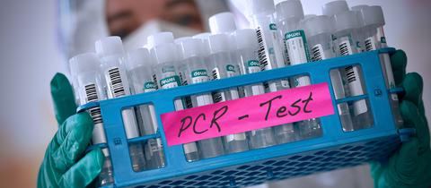 Foto einer medizinischen Fachangestellten in Schutzkleidung, die einen Korb mit sehr vielen Teströhrchen trägt, auf welchem der Aufkleber "PCR-Tests" angebracht ist.