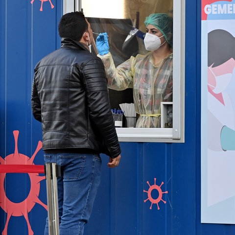 Ein Mann steht vor einem blauen Test-Container und wird durch ein Fenster hindurch von einer medizinischen Angestellten in Schutzkleidung mit einem Stäbchen in der Nase getestet.