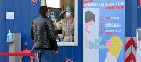 Ein Mann steht vor einem blauen Test-Container und wird durch ein Fenster hindurch von einer medizinischen Angestellten in Schutzkleidung mit einem Stäbchen in der Nase getestet.