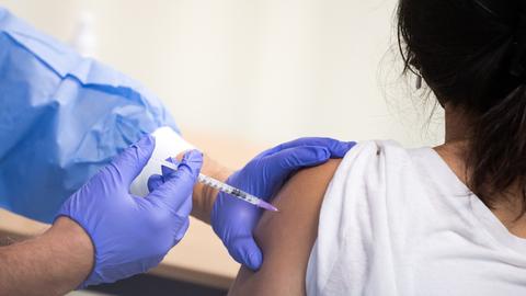 Hände mit blauen Schutzhandschuhen geben eine Spritze in den Oberarm einer Frau.