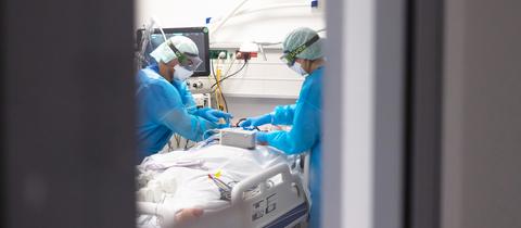 Ein Arzt und eine Pflegekraft in Schutzkleidung beugen sich über einen Patienten, der in einem Bett auf der Intensivstation liegt. Das Bild ist durch eine offene Tür fotografiert, weshalb das Bild rechts und links graue Flächen zeigt.