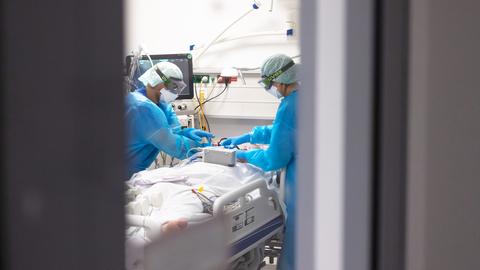 Ein Arzt und eine Pflegekraft in Schutzkleidung beugen sich über einen Patienten, der in einem Bett auf der Intensivstation liegt. Das Bild ist durch eine offene Tür fotografiert, weshalb das Bild rechts und links graue Flächen zeigt.