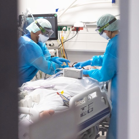 Ein Arzt und eine Pflegekraft in Schutzkleidung beugen sich über einen Patienten, der in einem Bett auf der Intensivstation liegt. Das Bild ist durch eine offene Tür fotografiert, weshalb das Bild rechts und links graue Flächen zeigt.