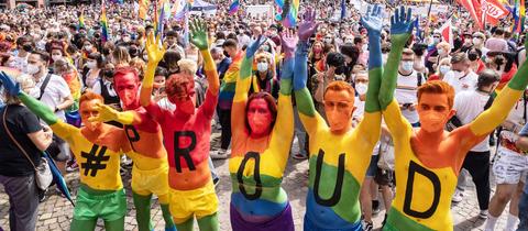 Sechs Menschena haben ihr Körper in Regenbogenfarben angemalt. Jeder der Körper trägt ein Zeichen: #,P,R,O,U,D. Sie recken ihre Arme nach oben. Im Hintergrund eine Masse von Menschen vor einer Fachwerkkulisse (Römer).