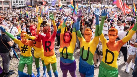 Sechs Menschena haben ihr Körper in Regenbogenfarben angemalt. Jeder der Körper trägt ein Zeichen: #,P,R,O,U,D. Sie recken ihre Arme nach oben. Im Hintergrund eine Masse von Menschen vor einer Fachwerkkulisse (Römer).