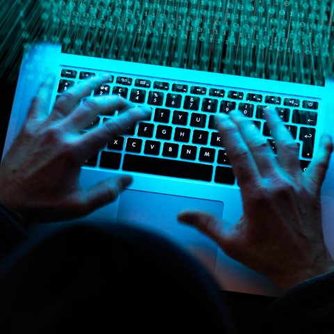 Foto von zwei Händen an einem Laptop, auf dessen Bildschirm viele O-en und 1-en leuchten. Die Umgebung ist dunkel.