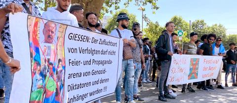 Menschen stehen nebeneinander in Reihe und halten zwei Banner hoch. Auf einem steht: "Gießen: Der Zufluchtsort von Verfolgten darf nicht Feier- und Propagandaarena von Genozid-Diktatoren und deren Anhänger werden".