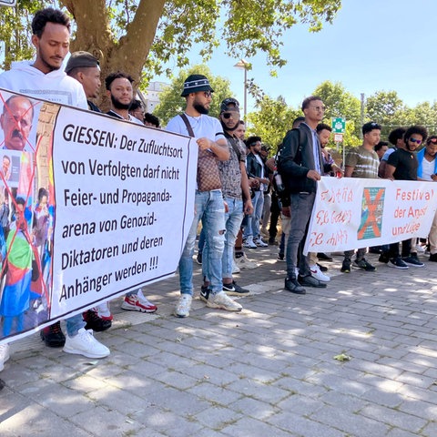 Menschen stehen nebeneinander in Reihe und halten zwei Banner hoch. Auf einem steht: "Gießen: Der Zufluchtsort von Verfolgten darf nicht Feier- und Propagandaarena von Genozid-Diktatoren und deren Anhänger werden".