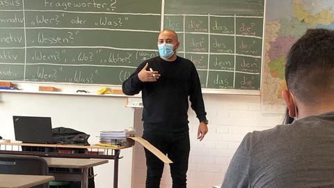 Ein Mann steht vor einer Tafel in einem Klassenraum. Die Tafel ist beschrieben mit Inhalten, welche deutsche Grammatik erklären.