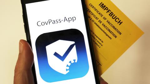 Das Logo der CovPass-App ist neben einem Impfbuch auf einem Smartphone zu sehen.