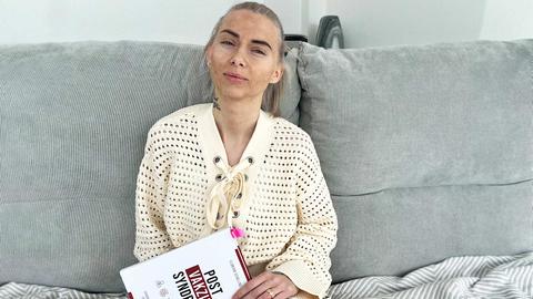 Eine Frau sitzt auf dem Sofa und hält ein Buch mit dem Titel "Post-Vac" in der Hand.