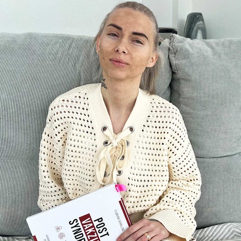 Eine Frau sitzt auf dem Sofa und hält ein Buch mit dem Titel "Post-Vac" in der Hand.