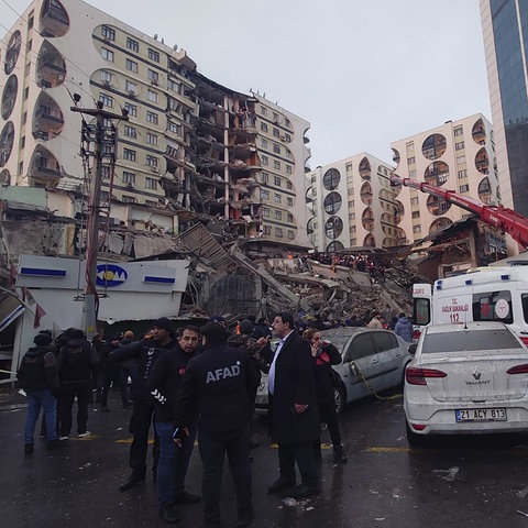 Menschen stehen am Boden, dahinter ein eingestützes Gebäude und ein roter Kran, der Gebäudeteile versucht anzuheben. 