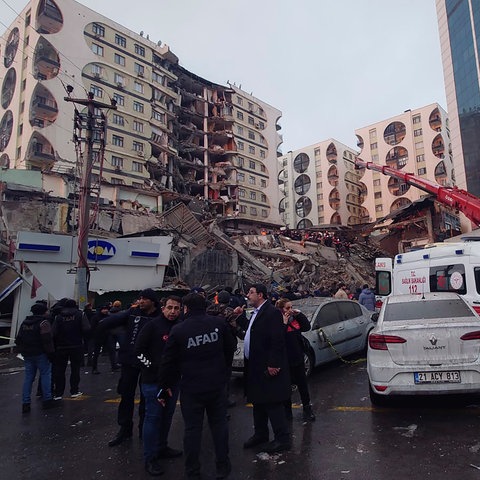 People stand on the ground, behind them a collapsed building and a red crane trying to lift parts of the building. 