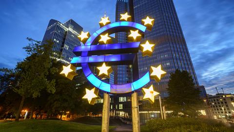 Das große Euro-Symbol vor der ehemaligen Zentrale der Europäischen Zentralbank (EZB, r) leuchtet am frühen Morgen.