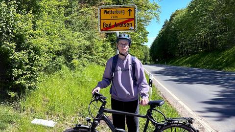 EIn junger Mann steht mit einem Fahrrad und Helm vor dem Ortsschild "Wetterburg 2 Km, Bad Aarolsen (durchgestrichen)". Im Hintergund die Landstraße mit Gebüsch rechts und links.
