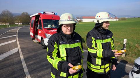 Zwei Feuerwehrleute mit Messgeräten in der Hand laufen über die Landstraße, ein Ferewehrauto im Hintergrund