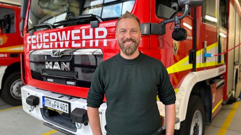 Tobias Hollomotz lehnt an der Front eines Feuerwehrautos. Hollomotz, Vollbart, lacht in die Kamera, Hände in den Hosentaschen, schwarzes Langarmshirt nach oben gekrempelt. 