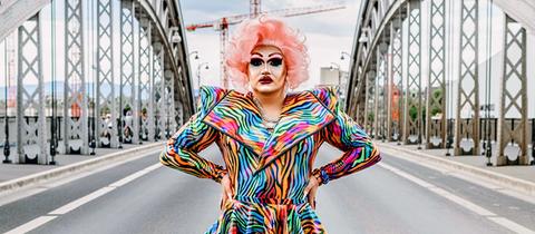 Dragqueen Electra Pain posiert in bundem Kleid und rosaner Perrücke auf einer Brücke.