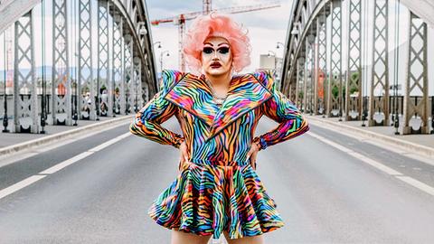 Dragqueen Electra Pain posiert in bundem Kleid und rosaner Perrücke auf einer Brücke.