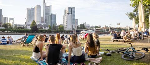 Menschen sitzen auf einer Rasenfläche am Fluss zusammen - mit Blick auf die Frankfurter Hochhäuser.