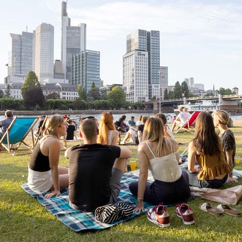 Menschen sitzen auf einer Rasenfläche am Fluss zusammen - mit Blick auf die Frankfurter Hochhäuser.