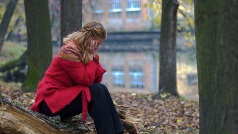 Frau, die in einem Stadtwald zusammengekauert auf den Boden sitzt und vor sich hin starrt.