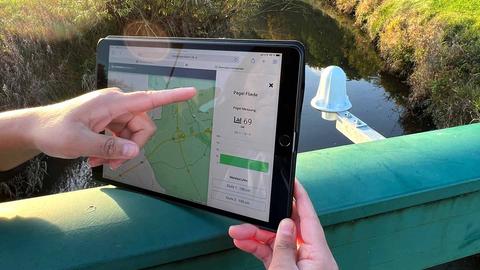 Hände halten ein Tablet an einen Sensor, der an einer Brücke angebracht ist. Auf dem Screen steht "Pegel Fiede". Im Hintergrund ein Bach neben einer Wiese.