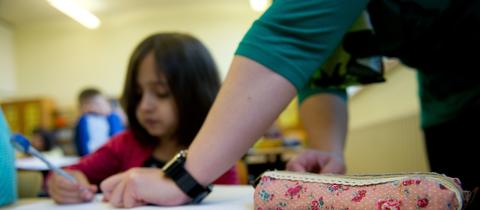 Szene in einer Schulklasse - ein Mädchen sitzt schreibend an einem Tisch - eine Hand stützt sich neben ihr auf. 