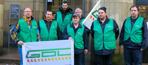 Sieben Männer in grünen GDL-Westen und einer GDL-Fahne stehen vor dem Eingang des Hauptbahnhofes Wiesbaden.