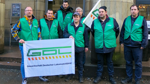Sieben Männer in grünen GDL-Westen und einer GDL-Fahne stehen vor dem Eingang des Hauptbahnhofes Wiesbaden.