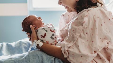 Eine Frau in einem Krankenhausbett hält einen Säugling vor sich in den Händen und lacht ihn an.