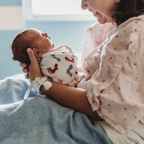 A woman in a hospital bed holds an infant in her hands in front of her and laughs at him.