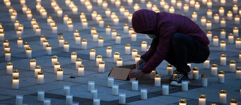 Sehr viele Grablichter stehen im Raster auf einem städtischen Boden. Die meisten leuchten, einige wenige nicht. Dazwischen eine Person, die in der Hocke sitzend ein Grablicht anzuündet.