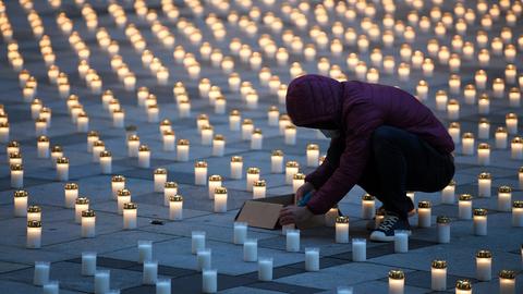 Sehr viele Grablichter stehen im Raster auf einem städtischen Boden. Die meisten leuchten, einige wenige nicht. Dazwischen eine Person, die in der Hocke sitzend ein Grablicht anzuündet.