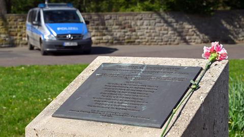 Eine Nelke liegt auf einem Gedenkstein, auf dem Namen eingraviert sind. Im Hintergrund steht ein Polizei-Streifenwagen.