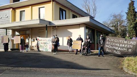 Foto einer Unterkunft für geflüchtete Menschen in Witzenhausen. Vor dem Gebäude stehen Menschen mit Schildern auf denen unter anderem steht: "Für guten und bezahlbaren Wohnraum! Für alle!" oder "Wie wollen bleiben"