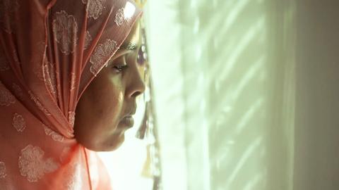 Nahaufnahme: Eine Frau mit einem Kopftuch steht an einem Fenster vor einem Vorhang und schaut mit leicht gesenktem Blick raus.