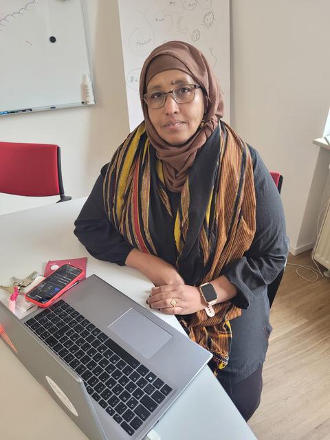 Hawo Abdulle, eine Frau mit Kopftuch, sitzt vor einem Computer an einem Schreibtisch und blickt in die Kamera