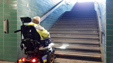 EIn Rollstuhlfahrer steht am Fuß einer Treppe, die nach oben zu einem Bahnsteig führt und blickt nach oben.