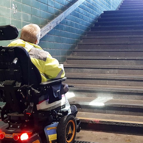 EIn Rollstuhlfahrer steht am Fuß einer Treppe, die nach oben zu einem Bahnsteig führt und blickt nach oben.