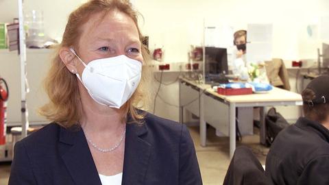 Eine Frau mit Mund-Nasen-Schutz steht vorne im Bild. Im Hintergrund sind Mitarbeitende an Schreibtischen und PCs zu sehen.