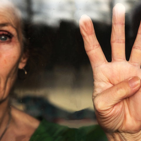 Eine Hand einer Frau, deren Gesicht zur Hälfte am Bildrand zu sehen ist. Hand und Frau befinden sich hinter einer Fensterscheibe - von außen betrachtet. Alle Finger der Hand sind ausgestreckt, der Daumen liegt im Handteller.