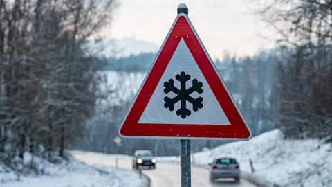 Ein Verkehrsschild mit einer Schneeflocke, das vor glatter Fahrbahn warnt steht am Straßenrand. 