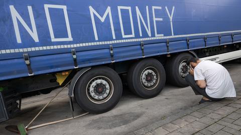 Ein Mann sitzt auf einer Raststätte vor einem Lkw, auf dessen Plane der Schriftzug "No Money" geklebt wurde.