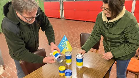 Zwei Menschen stehen mit Spendendosen in Ukrainefarben an einem Tisch in einer Feuerwehrhalle