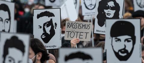 Nahaufnahme einer Demonstration. Viele Schilder mit den Portaits der Getöteten und in der Mitte eine Hand mit einem Schild, auf dem steht: "Rassismus tötet!".