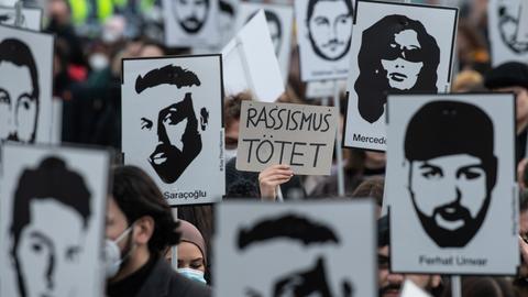 Nahaufnahme einer Demonstration. Viele Schilder mit den Portaits der Getöteten und in der Mitte eine Hand mit einem Schild, auf dem steht: "Rassismus tötet!".