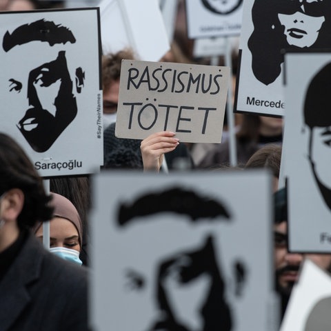 Nahaufnahme einer Demonstration. Viele Schilder mit den Portaits der Getöteten und in der Mitte eine Hand mit einem Schild, auf dem steht: "Rassismus tötet!".